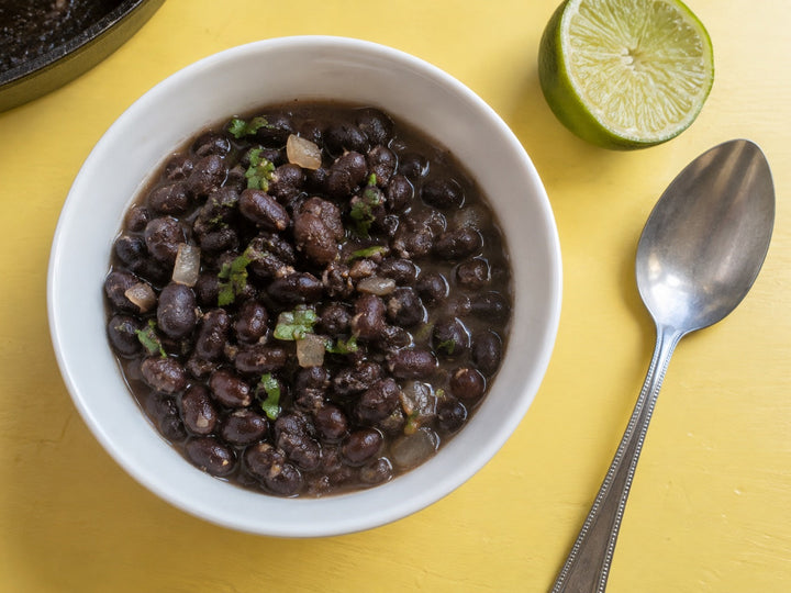 Black beans with onion and cilantro in a white bowl, with a lime half on a pale-yellow table.