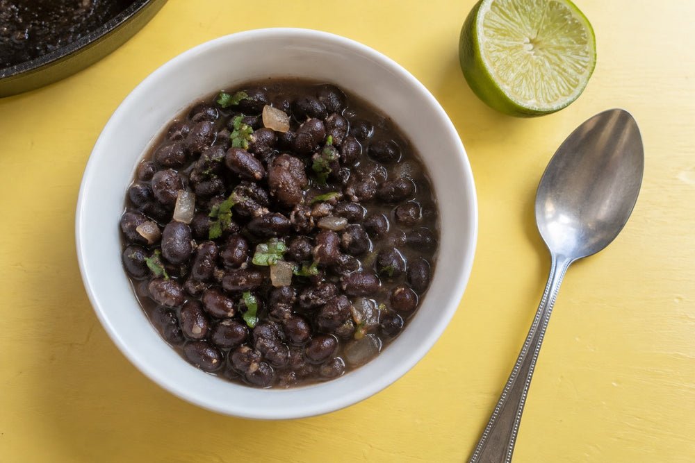 Black beans with onion and cilantro in a white bowl, with a lime half on a pale-yellow table.