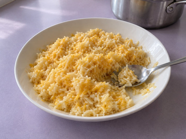 Cheddar-coated rice with distinct grains in a shallow white dish on a pale-lavender tabletop.