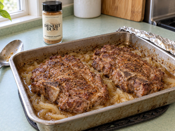 Two browned pork shoulder steaks braised with onion in a metal baking pan beside Original Desert Dust.