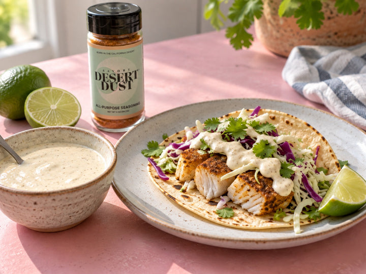 Creamy lime fish taco sauce in a bowl with a cooked fish taco as a serving suggestion. Savory Desert Dust jar nearby.