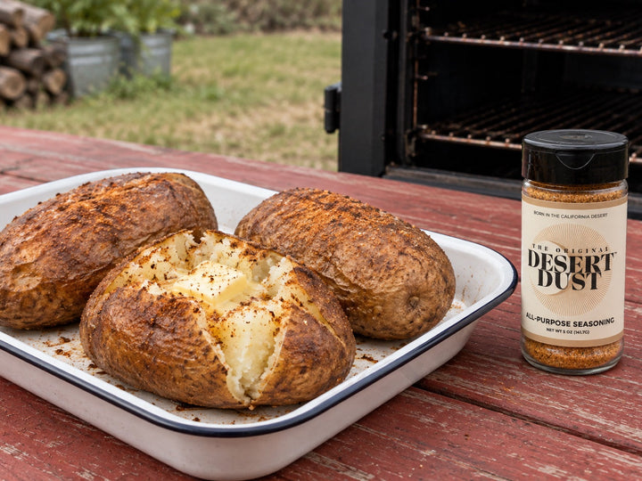 Split smoked russet potatoes with fluffy centers beside an Original Desert Dust jar and a backyard smoker.