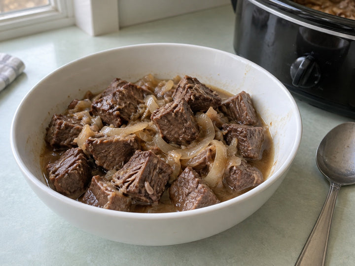 Tender braised chuck beef bites with onion and light brown gravy in a small white bowl beside a slow cooker.