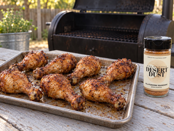 Browned smoked chicken drumsticks on a tray beside a backyard smoker and an Original Desert Dust jar.