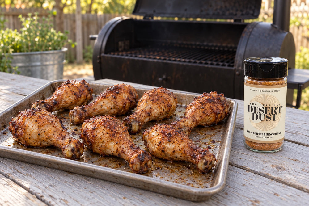 Browned smoked chicken drumsticks on a tray beside a backyard smoker and an Original Desert Dust jar.