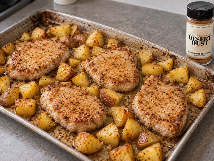 Oven-baked pork loin chops and golden potato pieces on a worn sheet pan beside an Original Desert Dust jar.