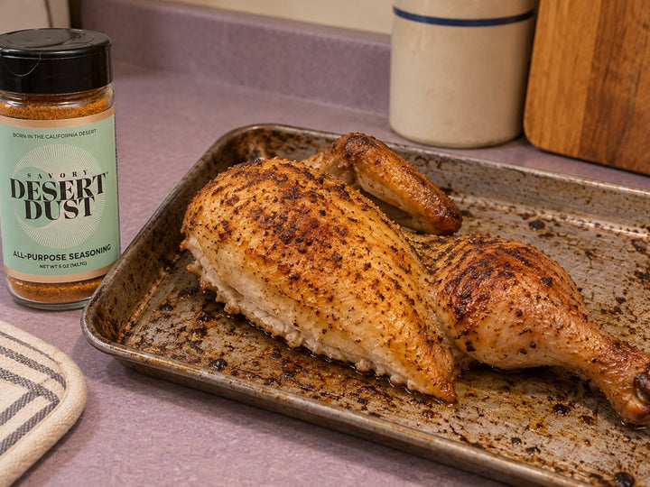 One roasted chicken half with a single breast, wing and leg on a small tray beside a Savory Desert Dust jar.