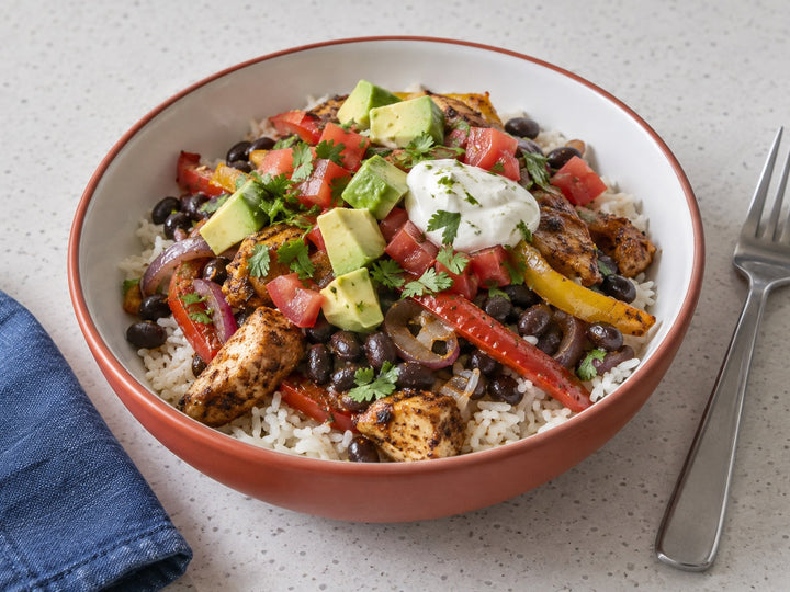 Chicken fajita bowl with white rice, black beans, peppers, red onion, avocado, tomato and a small lime-yogurt topping.