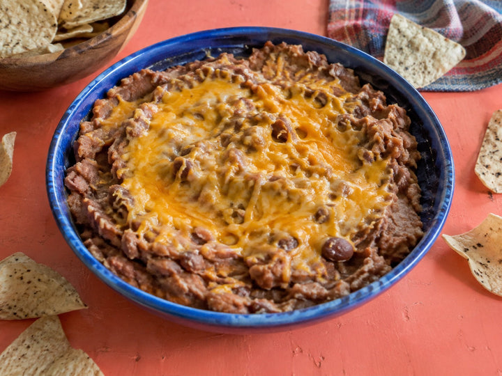 Warm mashed pinto bean dip with melted cheddar in a blue bowl beside tortilla chips.