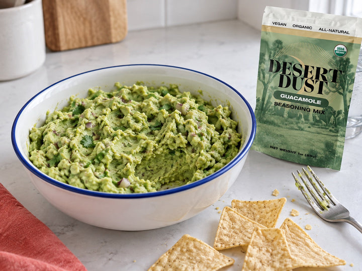 Chunky homemade guacamole in a blue-rimmed white bowl, with tortilla chips and a Desert Dust Guacamole Seasoning pouch.