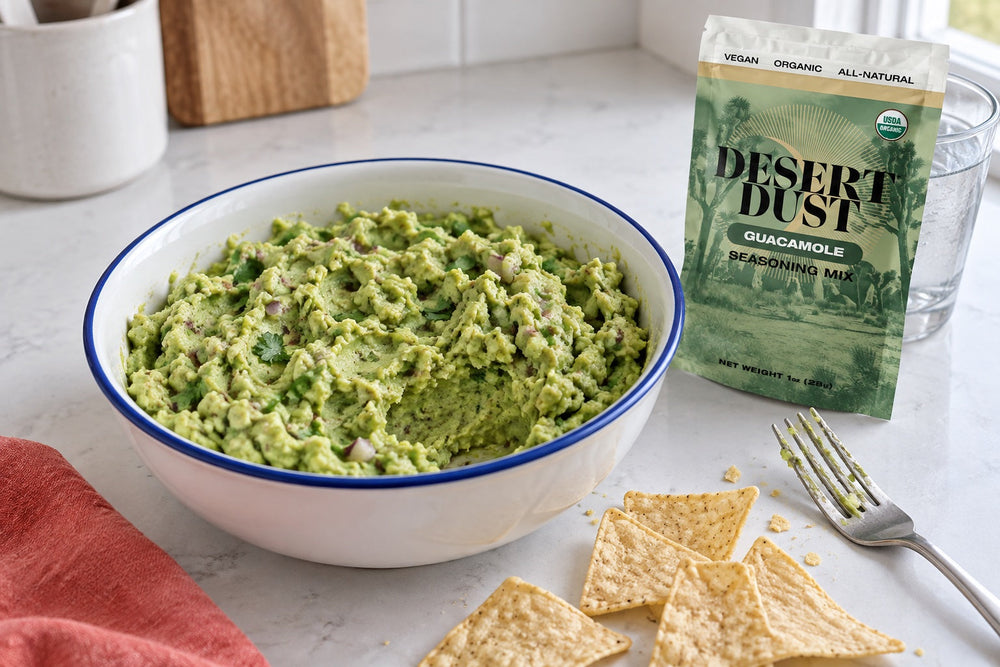 Chunky homemade guacamole in a blue-rimmed white bowl, with tortilla chips and a Desert Dust Guacamole Seasoning pouch.