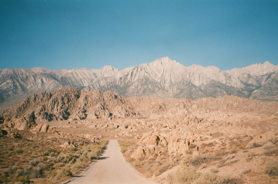 A pale dirt road crossing rocky desert terrain toward distant mountains.