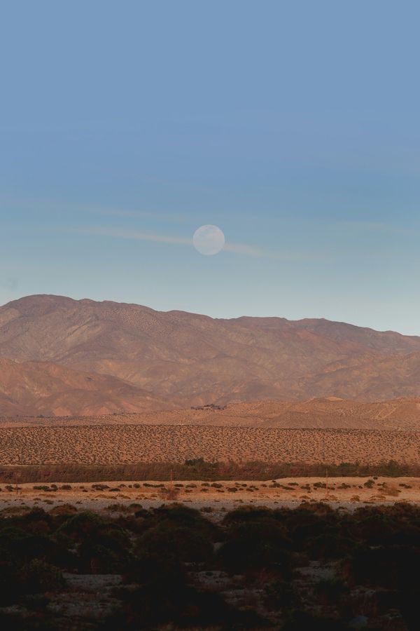 A pale moon rises above warm desert mountains and low shrubs.