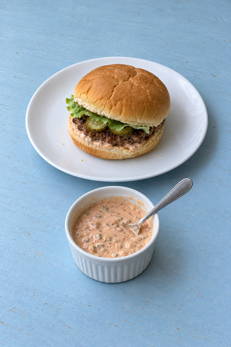 Burger sauce in a white ramekin beside a simple homemade burger on a white plate and blue kitchen table.