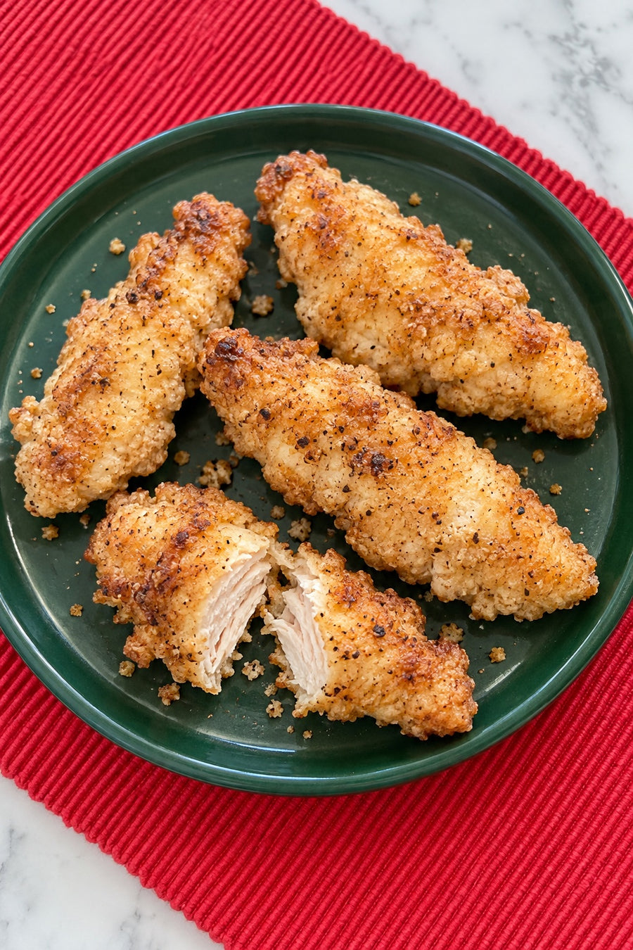 Golden flour-coated chicken tenders on a green plate over a red cloth, with one tender opened to show the cooked center.