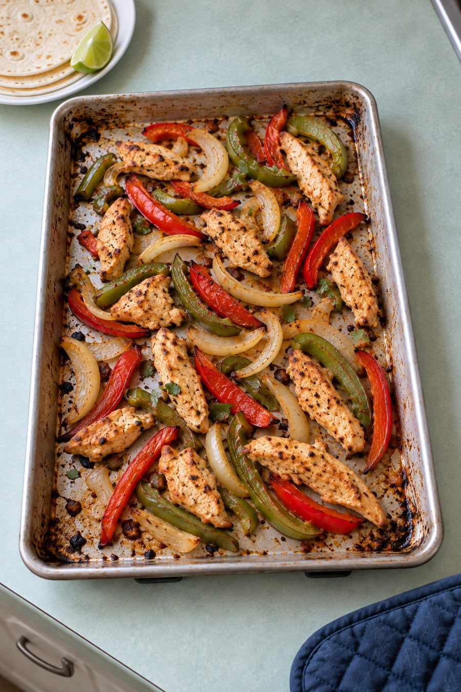A metal tray of oven chicken fajitas with red and green pepper strips, yellow onion and tortillas nearby on a mint counter.