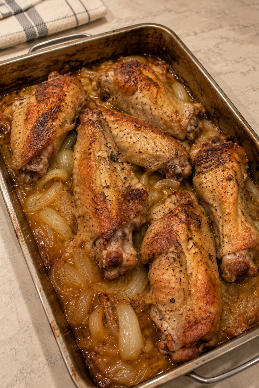 Baked turkey wing sections with browned skin, soft onions and cooking juices in a roasting pan.